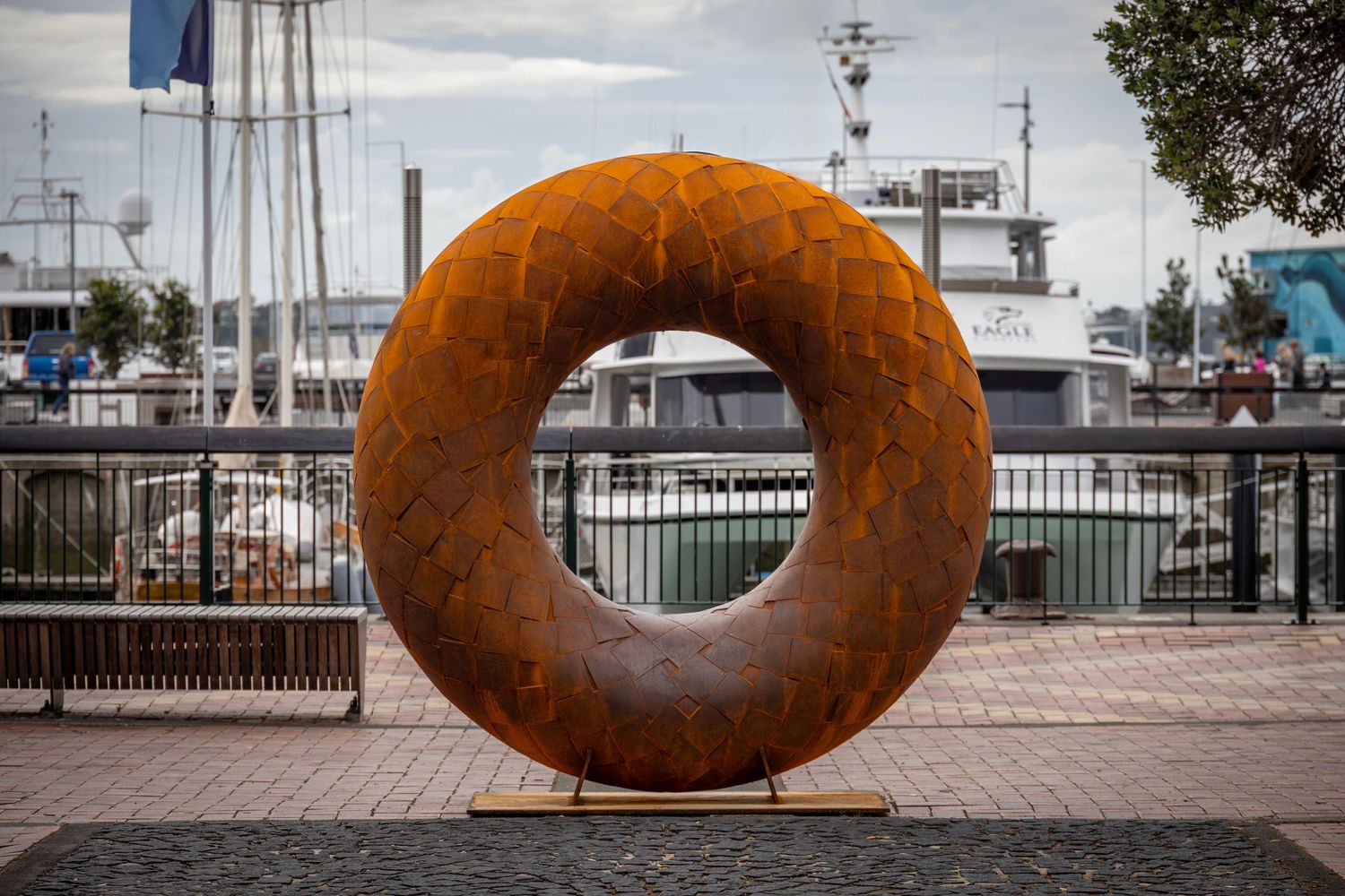 Circular Corten steel sculpture with a scaled texture, displayed on a waterfront promenade with boats and railings in the background.