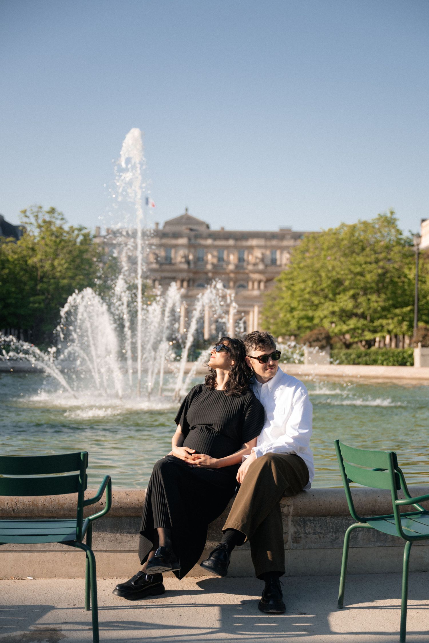 A couple wearing sunglasses relaxing together outdoors on a sunny day, with green foliage in the background.
