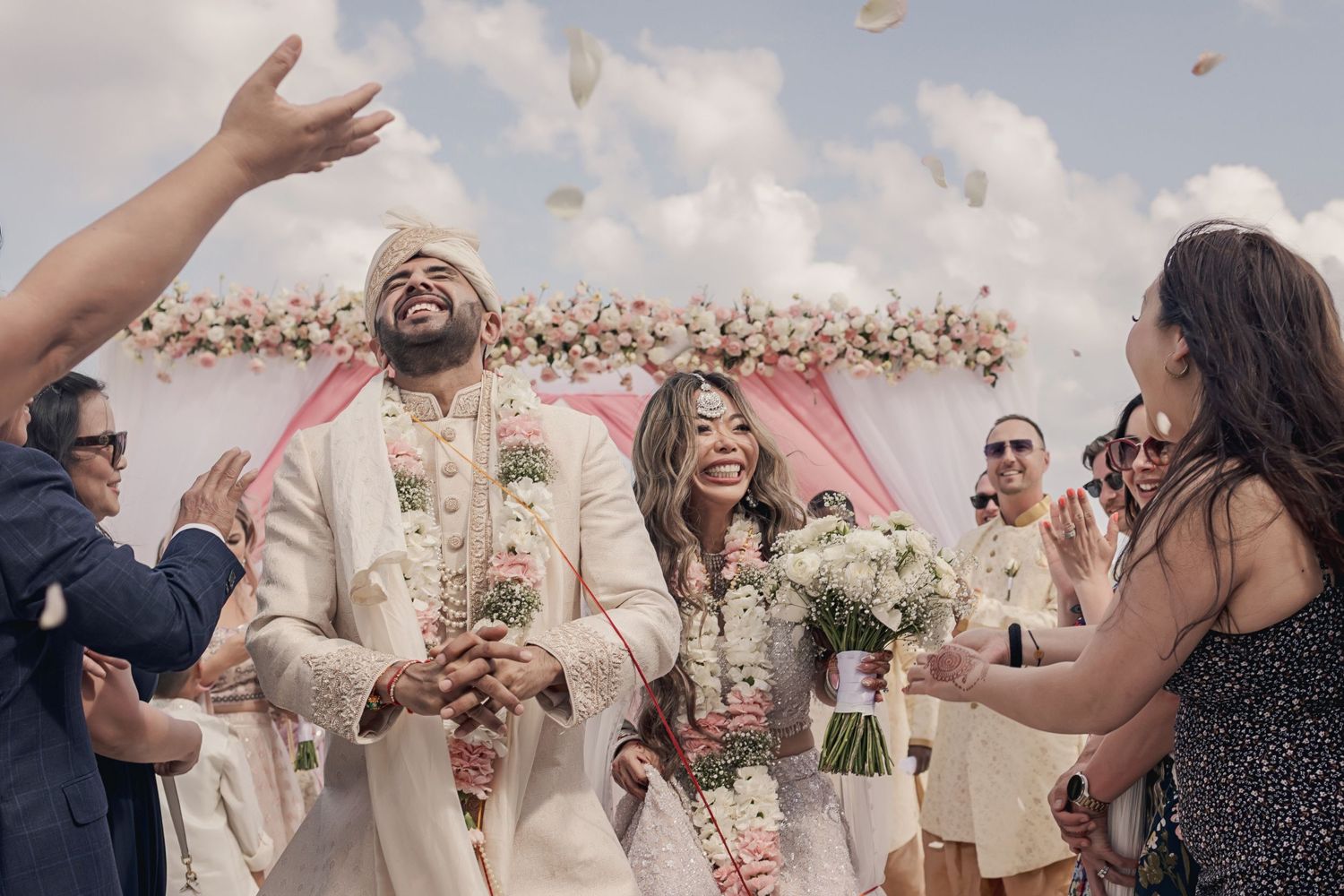 Wedding party celebrates joyfully with flower petals being tossed in the air at an outdoor beach ceremony.