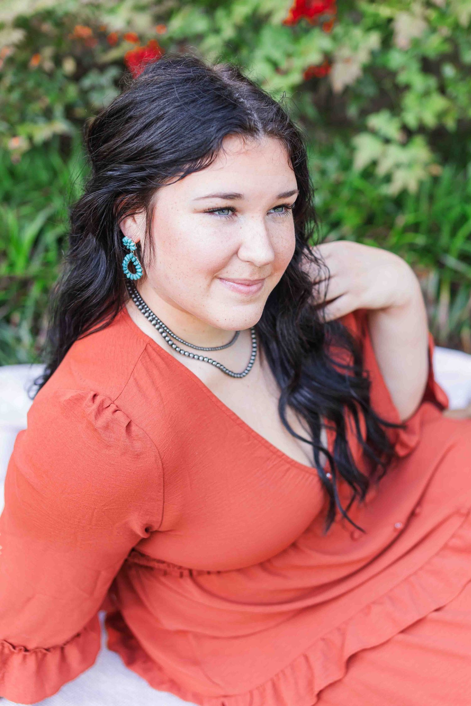 Woman in coral red dress with long dark hair and turquoise earrings poses outdoors near red flowers.