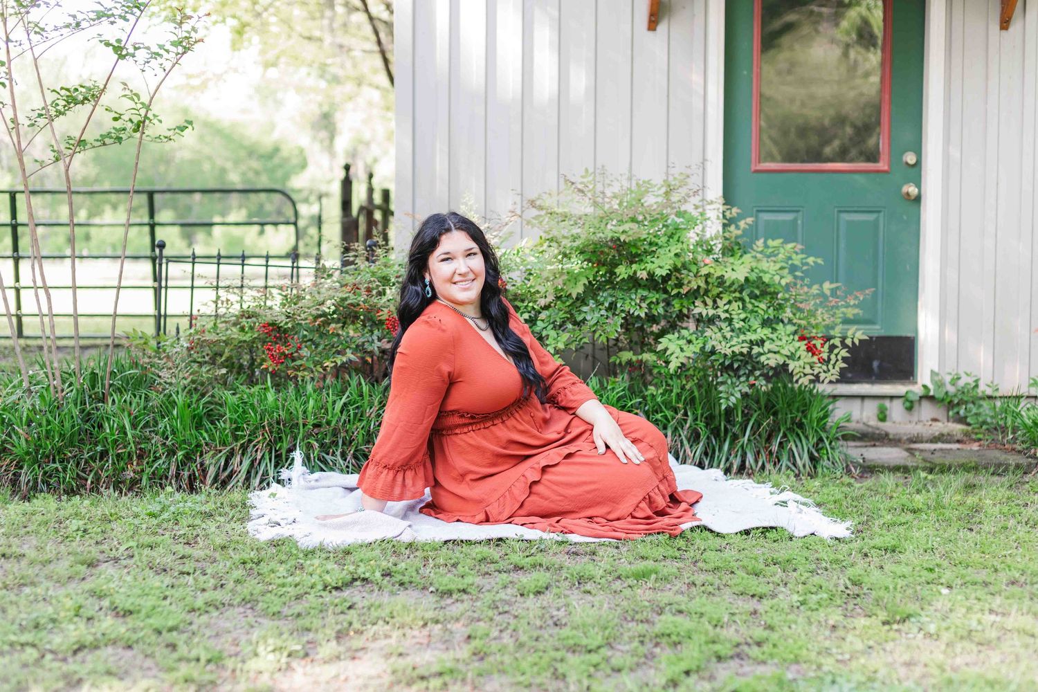 A person in a burnt orange dress sits on a white blanket outside a rustic home with green bushes and door.