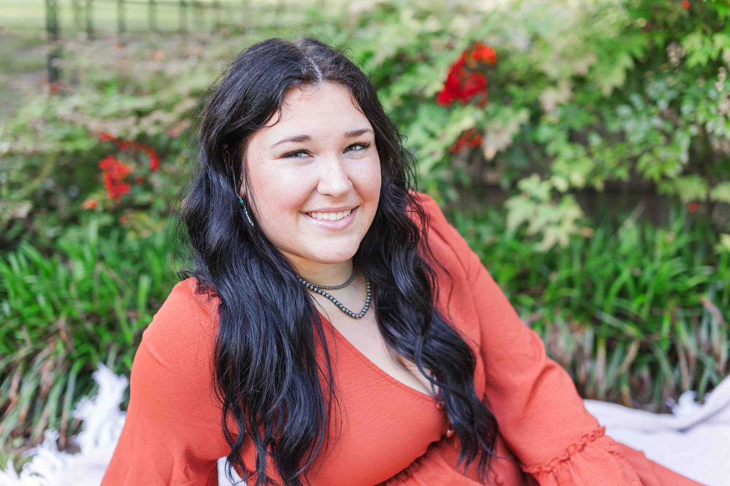 A smiling figure wearing a coral sweater sits in a garden with red flowers and greenery in the background.