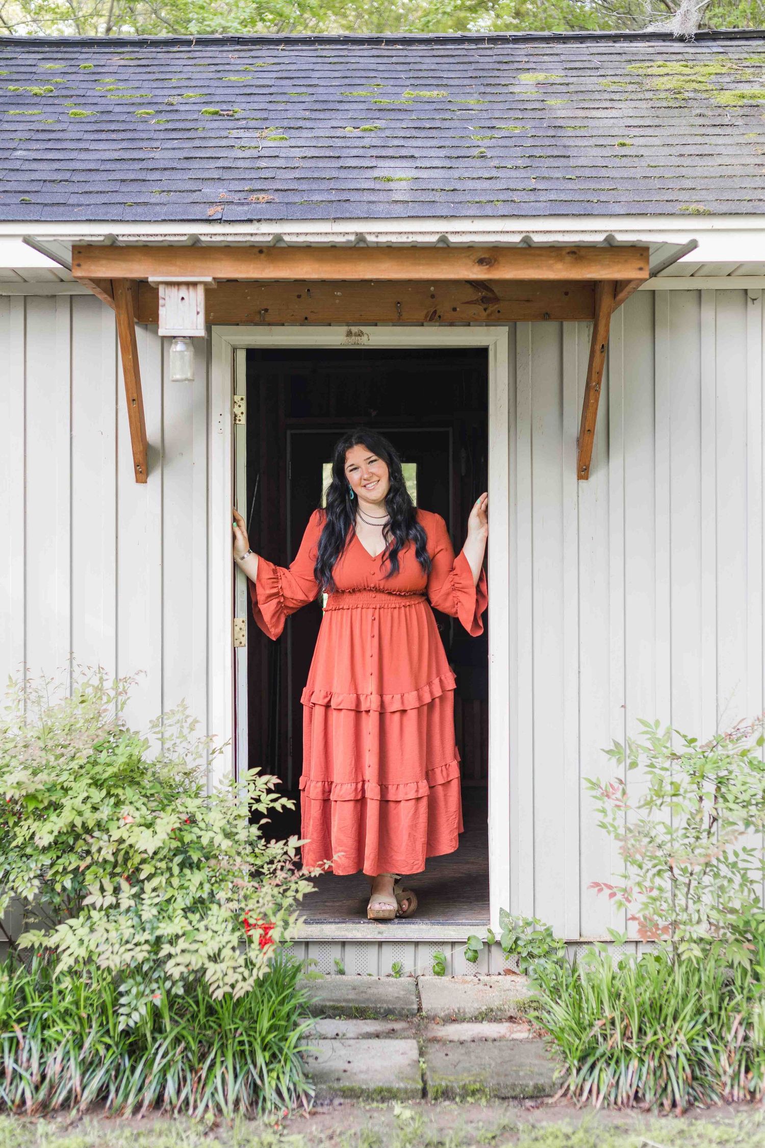 A rust-colored tiered maxi dress is showcased in a rustic white doorway with a garden in the foreground.