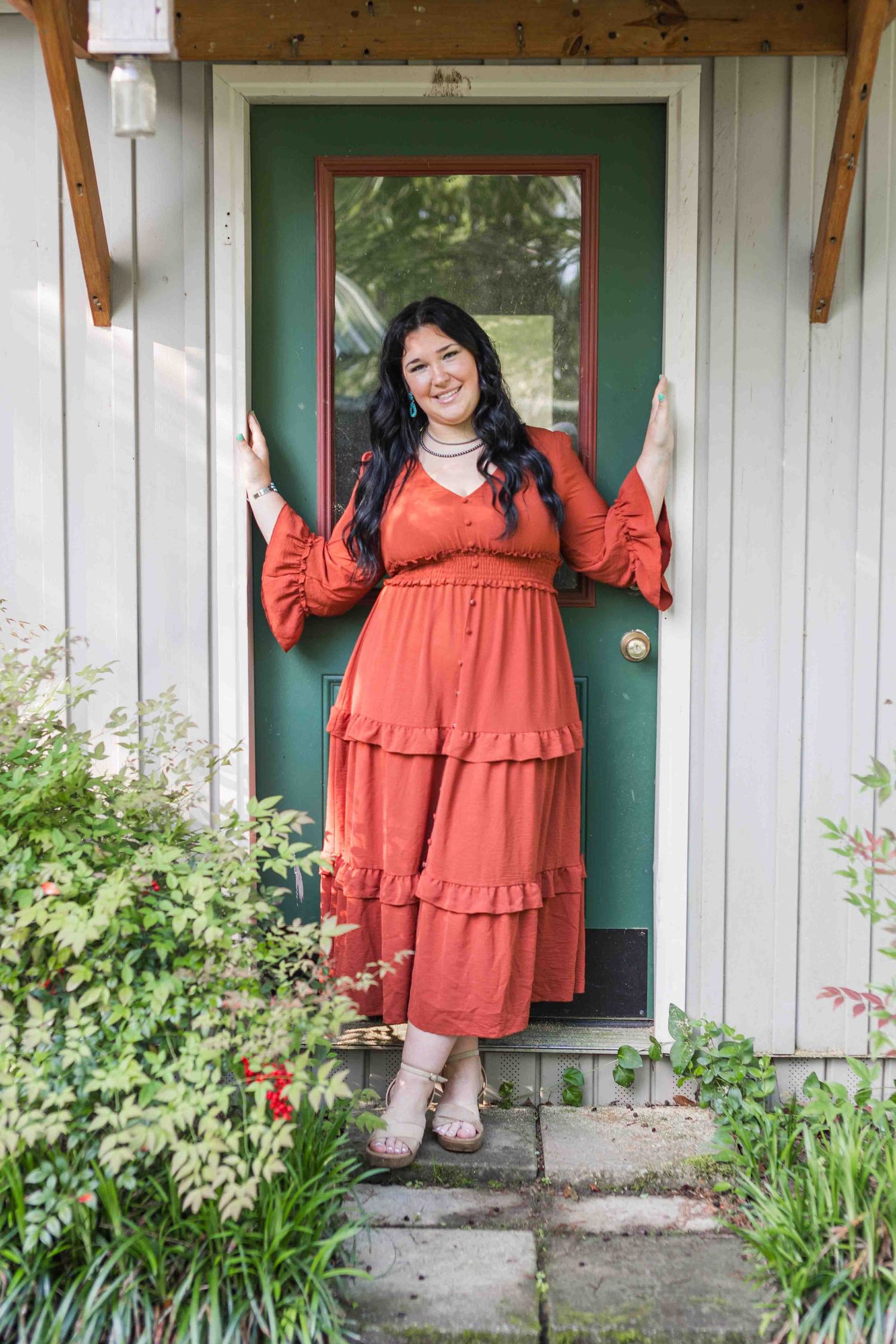 A flowing coral dress is featured against a turquoise door frame surrounded by garden flowers and greenery.