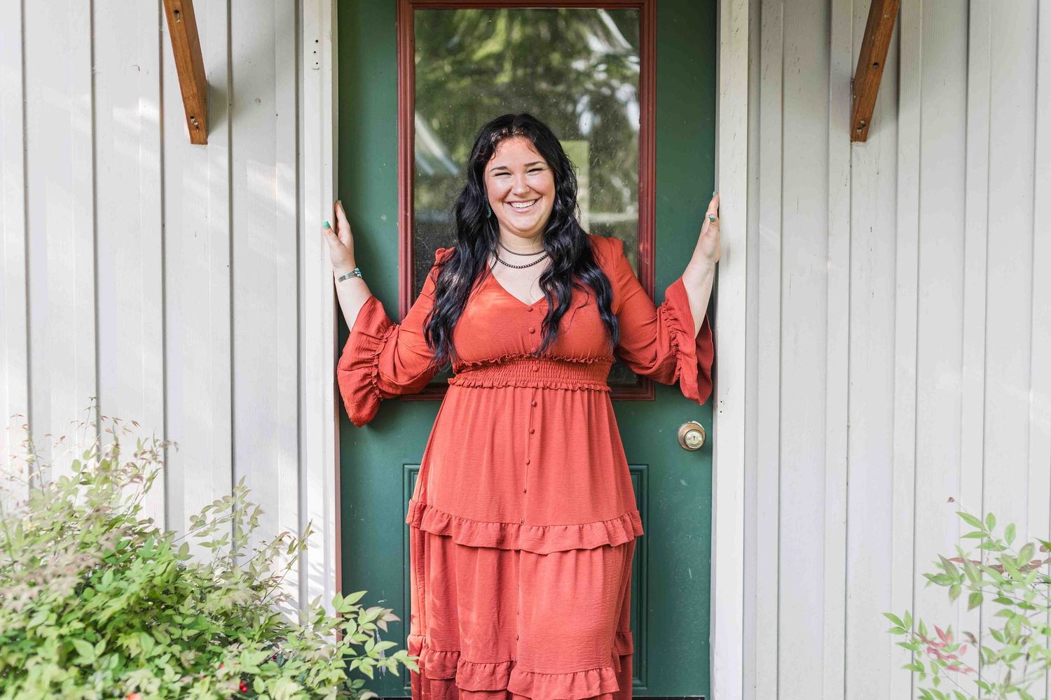 Someone wearing a coral tiered maxi dress stands smiling in a doorway with white trim and a green door.