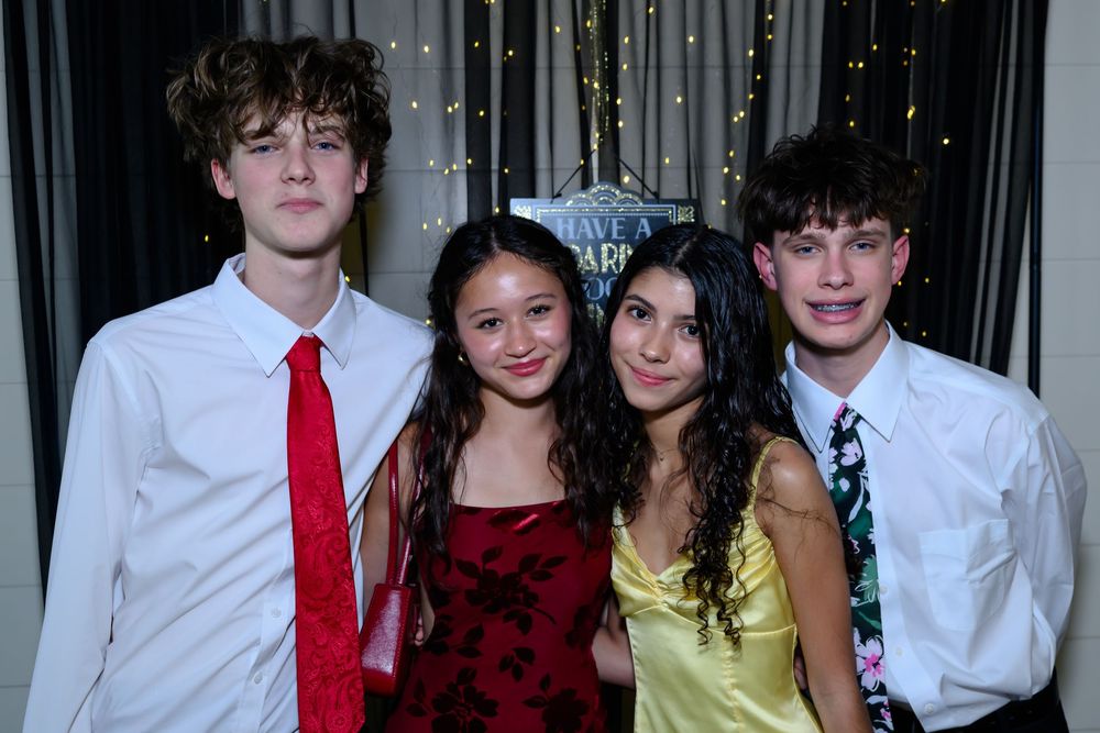 A group of four teens pose together at a formal event wearing dress clothes in front of sparkly curtains.