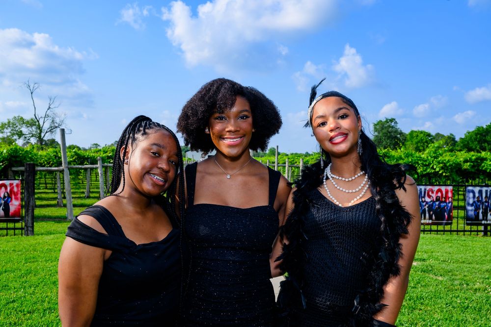 Three people wearing black outfits smile together on a sunny day at an outdoor venue with green grass and trees.