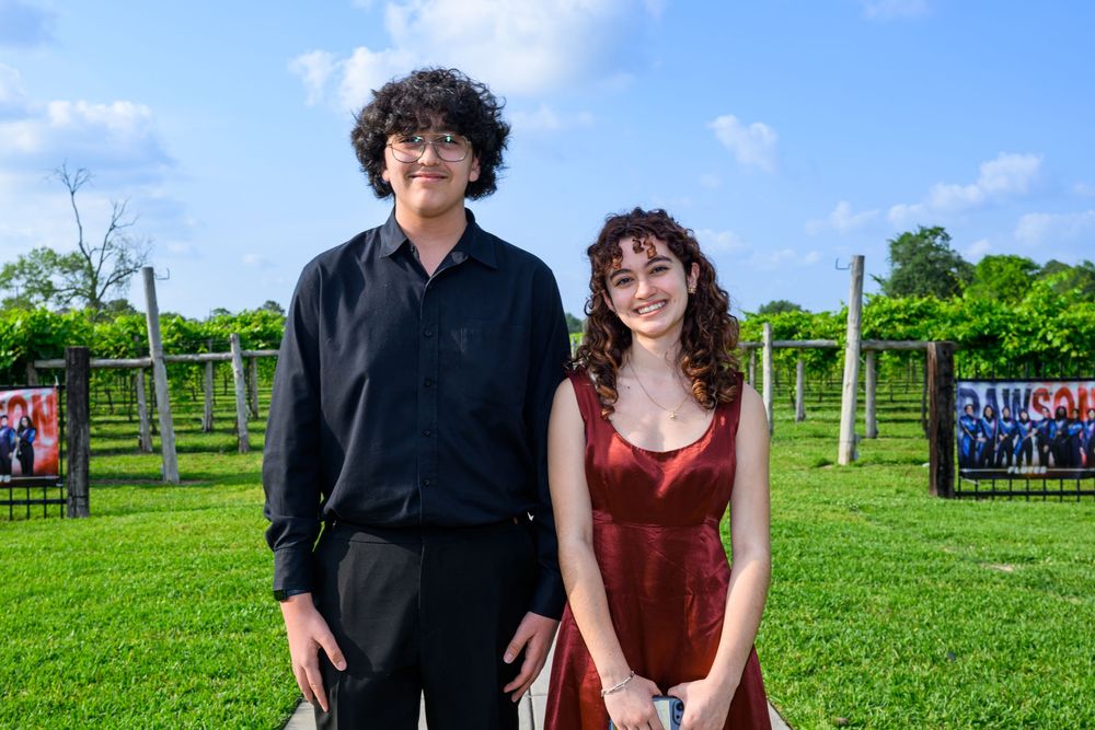 People in formal attire pose together outdoors on a bright green lawn with grapevine trellises in the background.