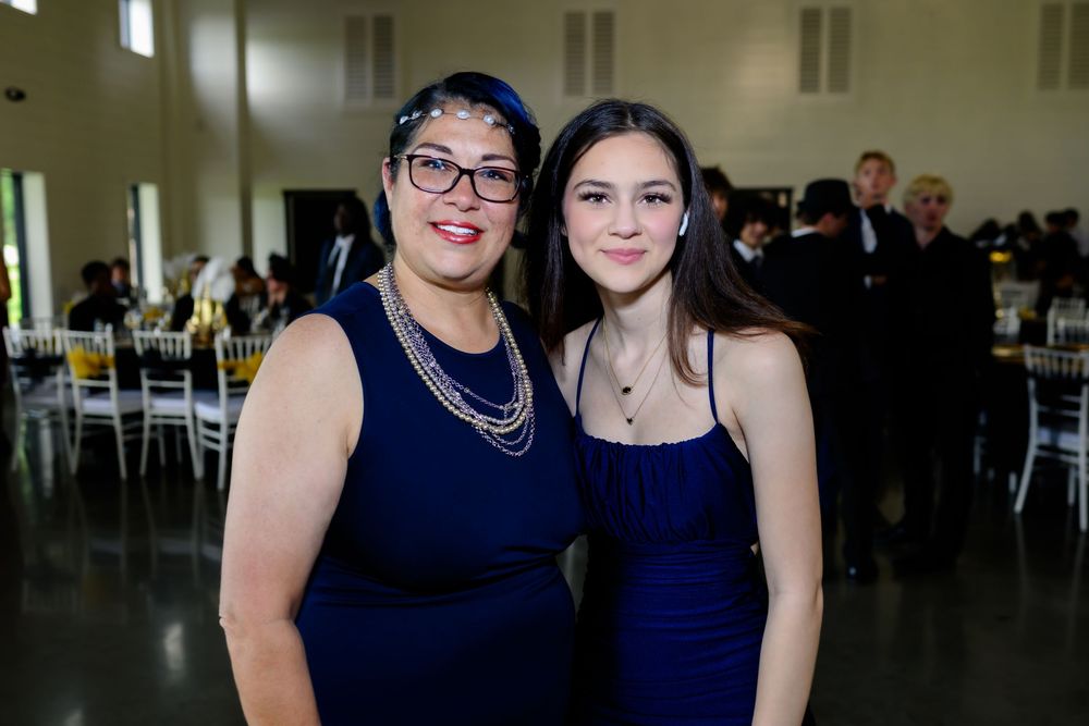 Two people in matching navy blue dresses pose together at a formal event with tables and chairs visible in background.