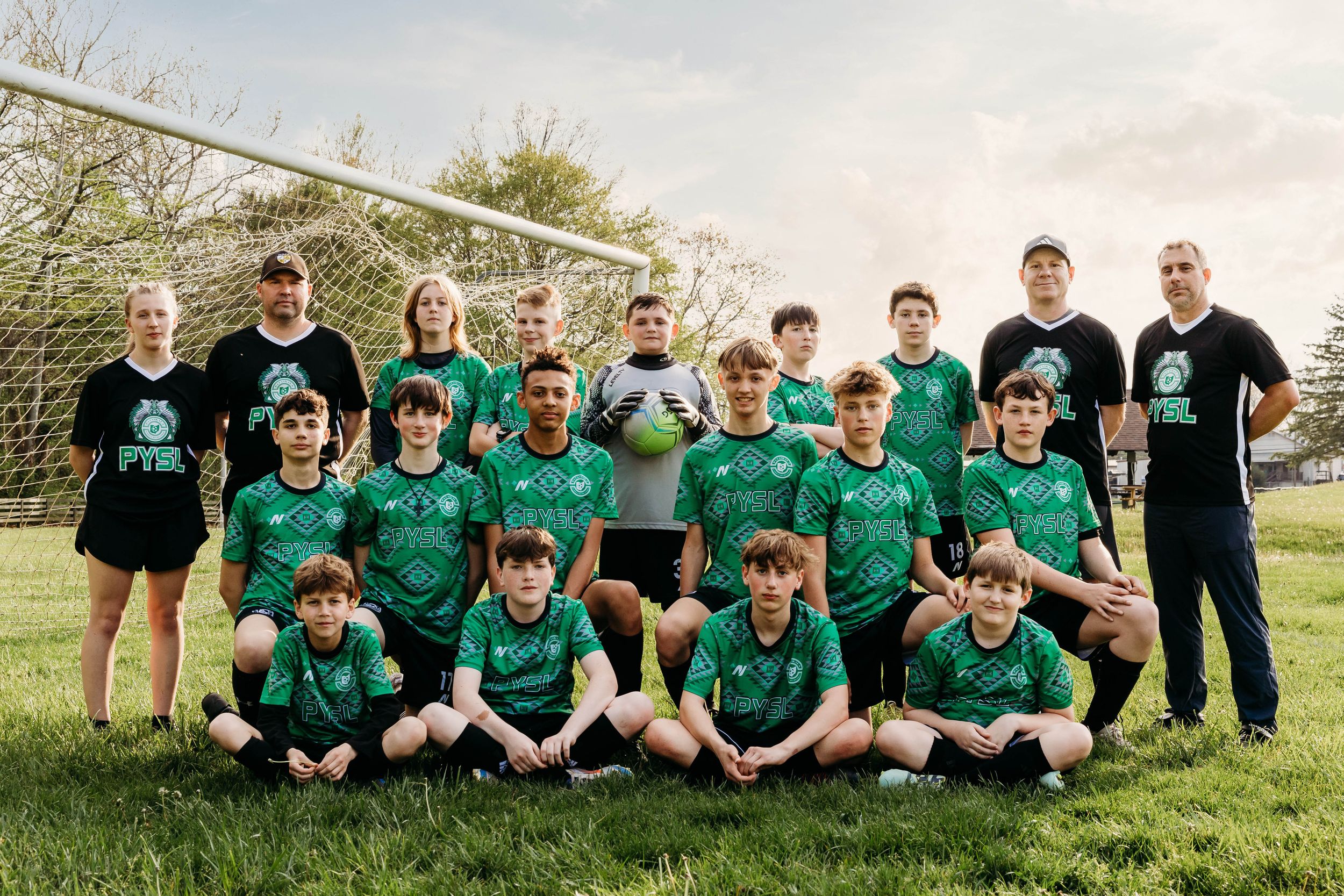 A youth soccer team in green jerseys and their coaches pose together on a grass field in front of a soccer goal.