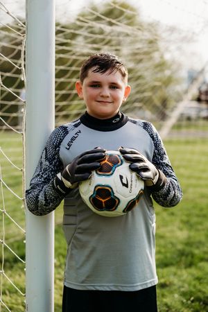 A young soccer goalkeeper in gray jersey and gloves holds a soccer ball while standing by the goal post on a grassy field.