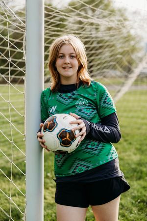 Soccer goalkeeper in a green patterned jersey holds a ball while standing by the goal post on a grassy field.