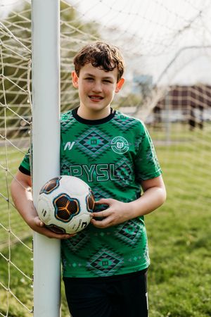Young soccer player in green patterned jersey holds ball while standing next to goal post on grass field.