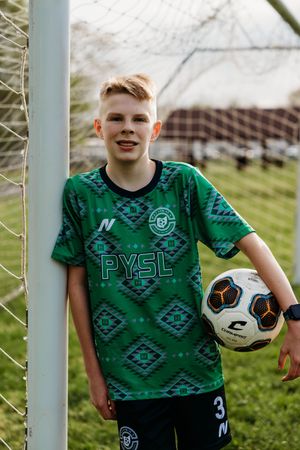 Soccer player stands by goal post wearing green patterned jersey while holding ball and smiling on grass field.