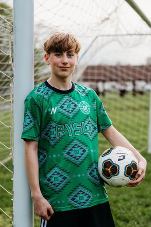 Soccer player wearing green patterned jersey stands by goal post holding white ball on grass field.