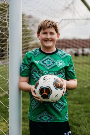 Soccer player in green patterned jersey holding a black and white ball next to goal post at field.