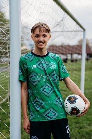 Soccer player wearing a green geometric patterned jersey holds a soccer ball while standing by a goal post on grass field.
