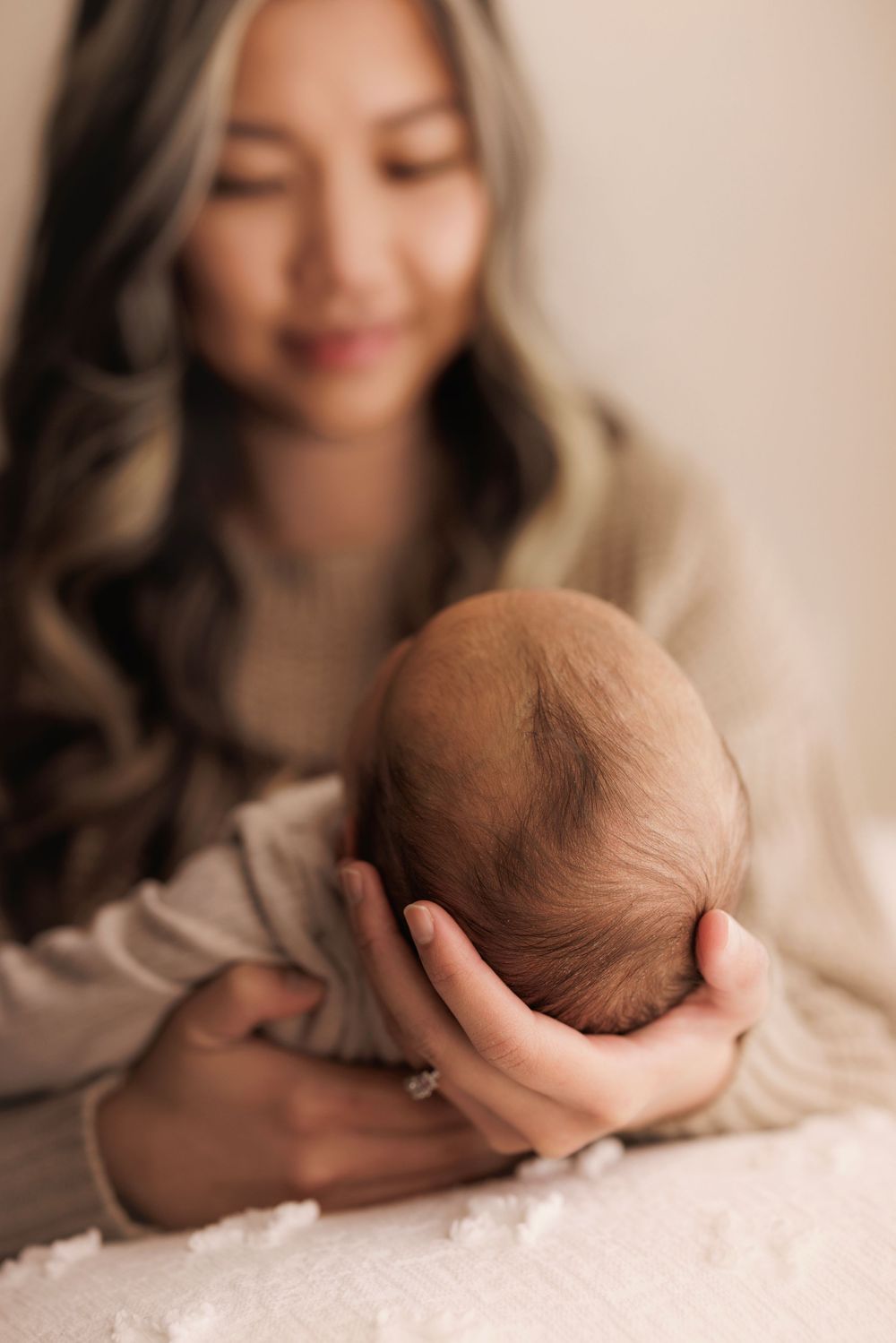 Gentle portrait of a mother in beige clothing holding her sleeping newborn baby against soft white bedding.