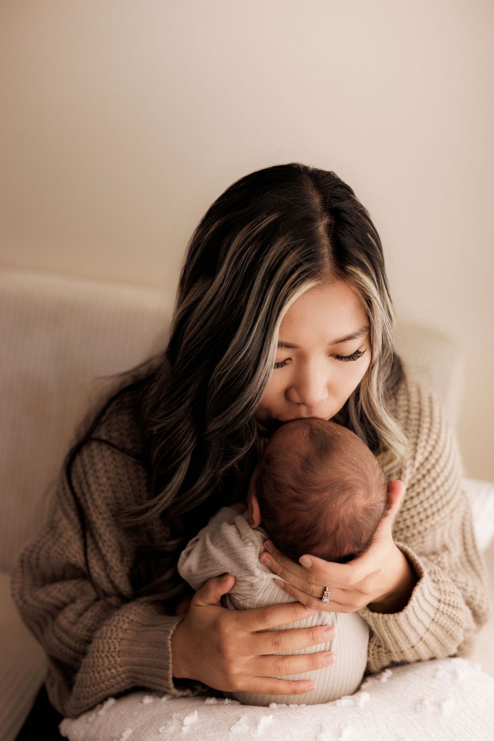 Woman in cozy beige sweater tenderly embraces and kisses newborn baby in intimate indoor portrait.