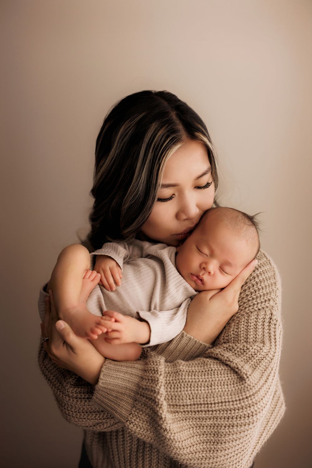 A tender moment captures a mother in a knit sweater embracing and kissing her sleeping newborn baby.