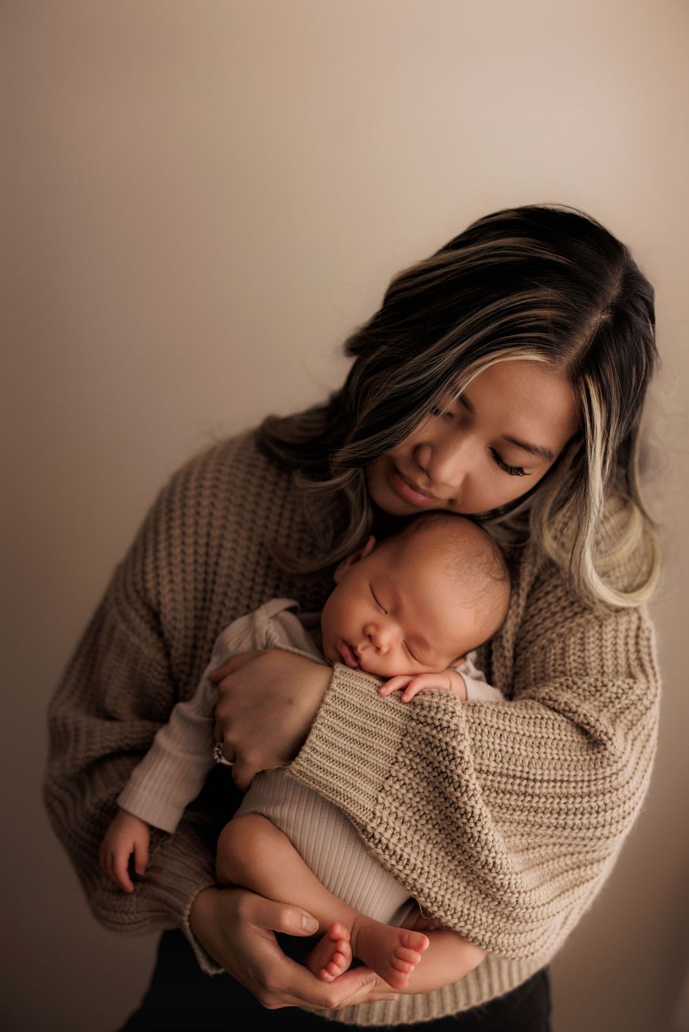 A tender moment captured between a mother and sleeping newborn baby in matching beige knit sweaters against neutral background.