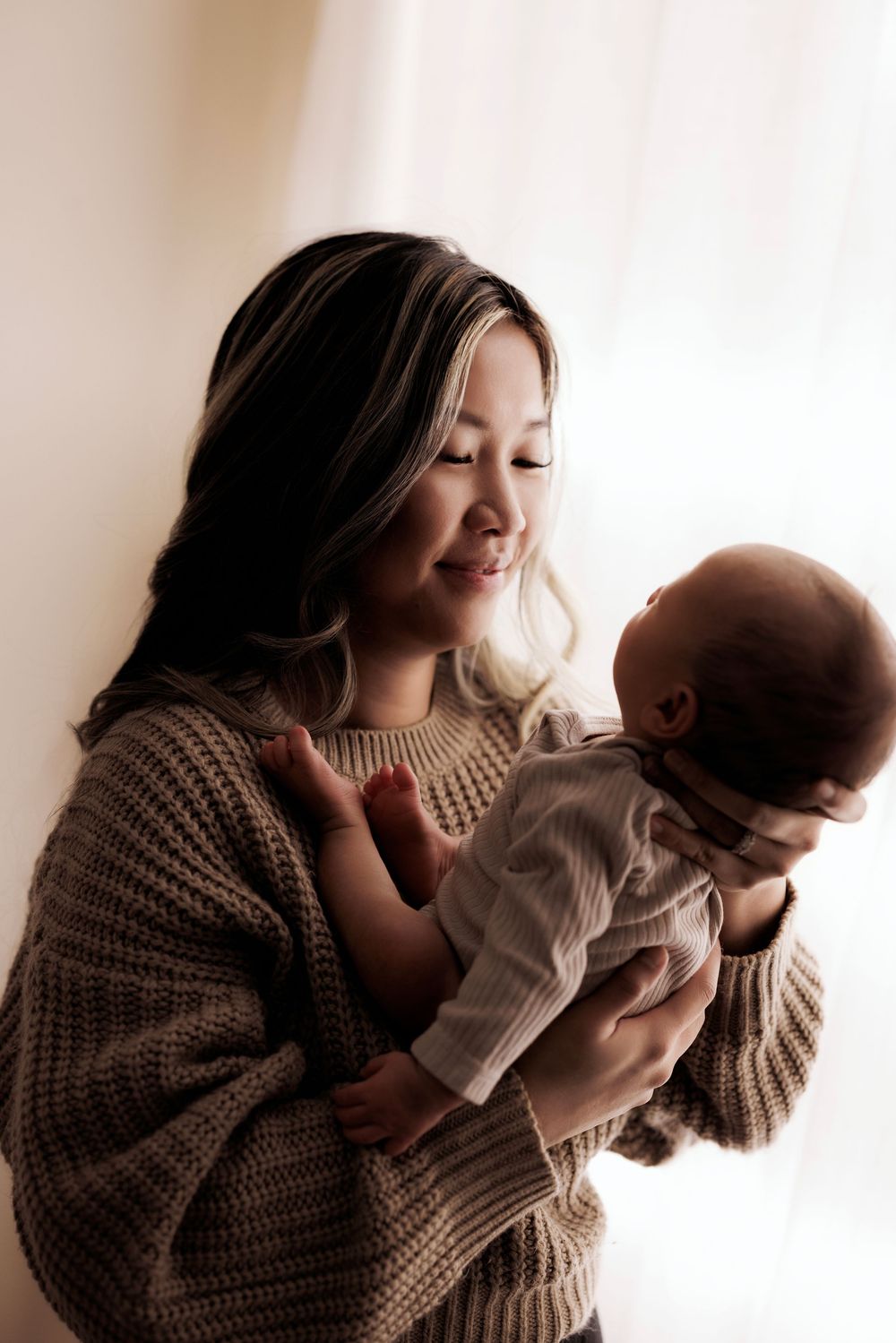 A mother in a cozy knit sweater holds and gazes lovingly at her baby near a sunlit window.