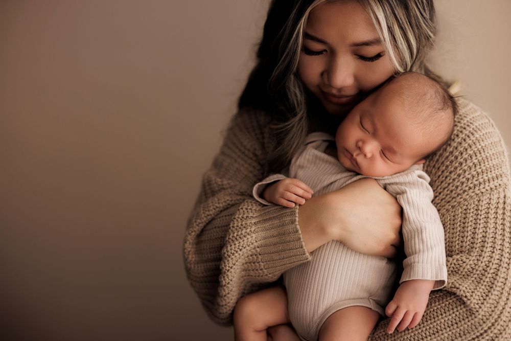 A tender moment as a baby sleeps peacefully in someone's arms while wearing a white onesie against a neutral backdrop.