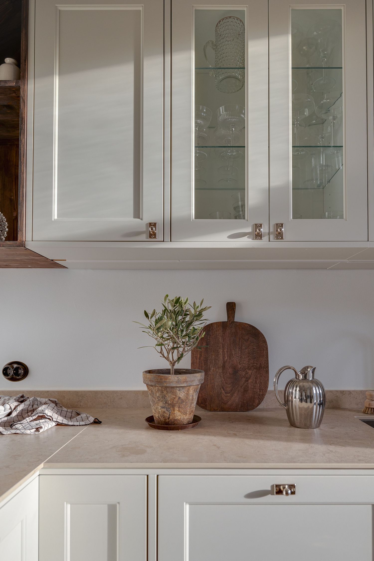 White kitchen cabinets with glass doors and rustic decor featuring a potted olive plant and wooden cutting board on marble counter.