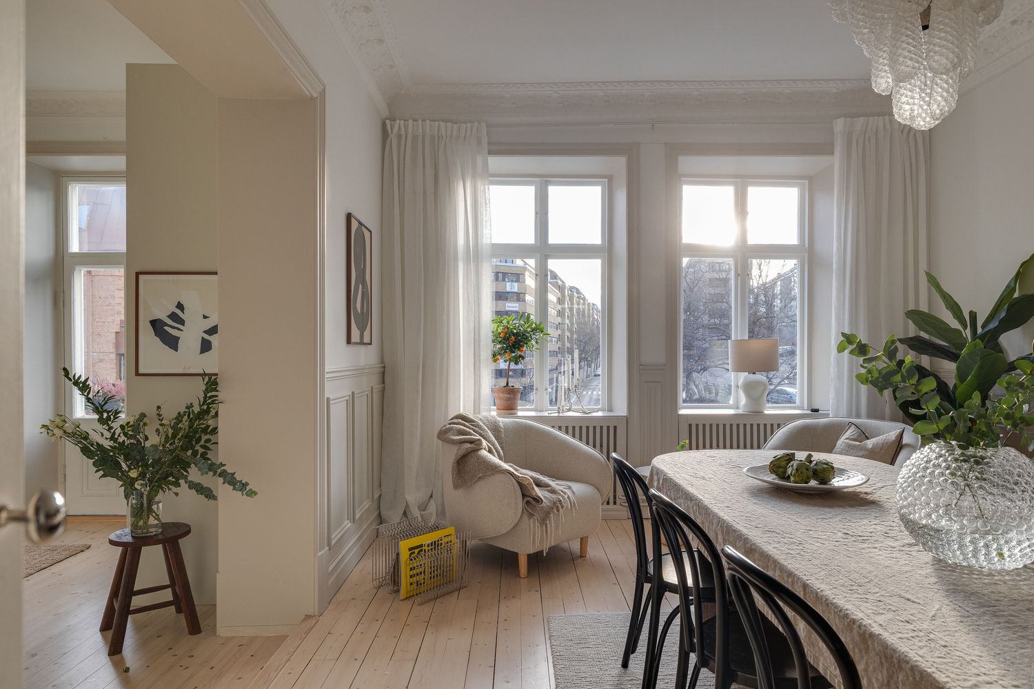 Bright Scandinavian dining room with wooden floors, large windows, hanging crystal lamp and indoor plants.
