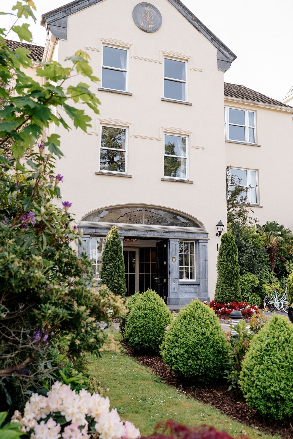 Elegant white country house with arched entrance and manicured garden featuring trimmed hedges and colorful flowers.