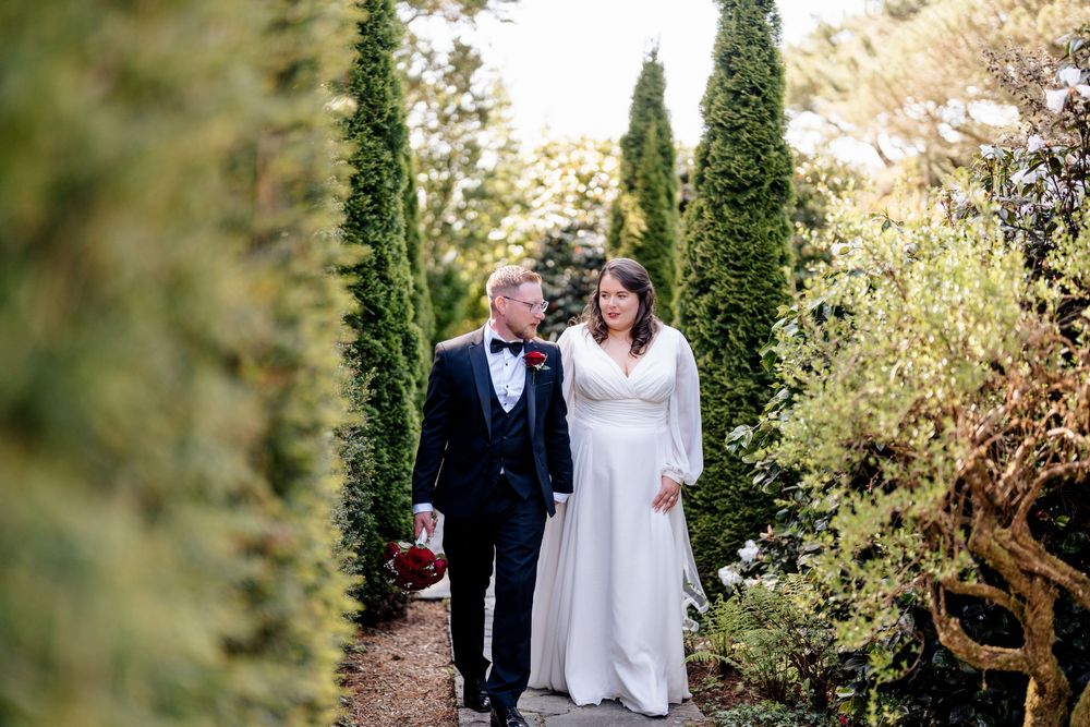 A couple on their wedding day walking along a garden path lined with tall cypress trees and greenery on a sunny day.