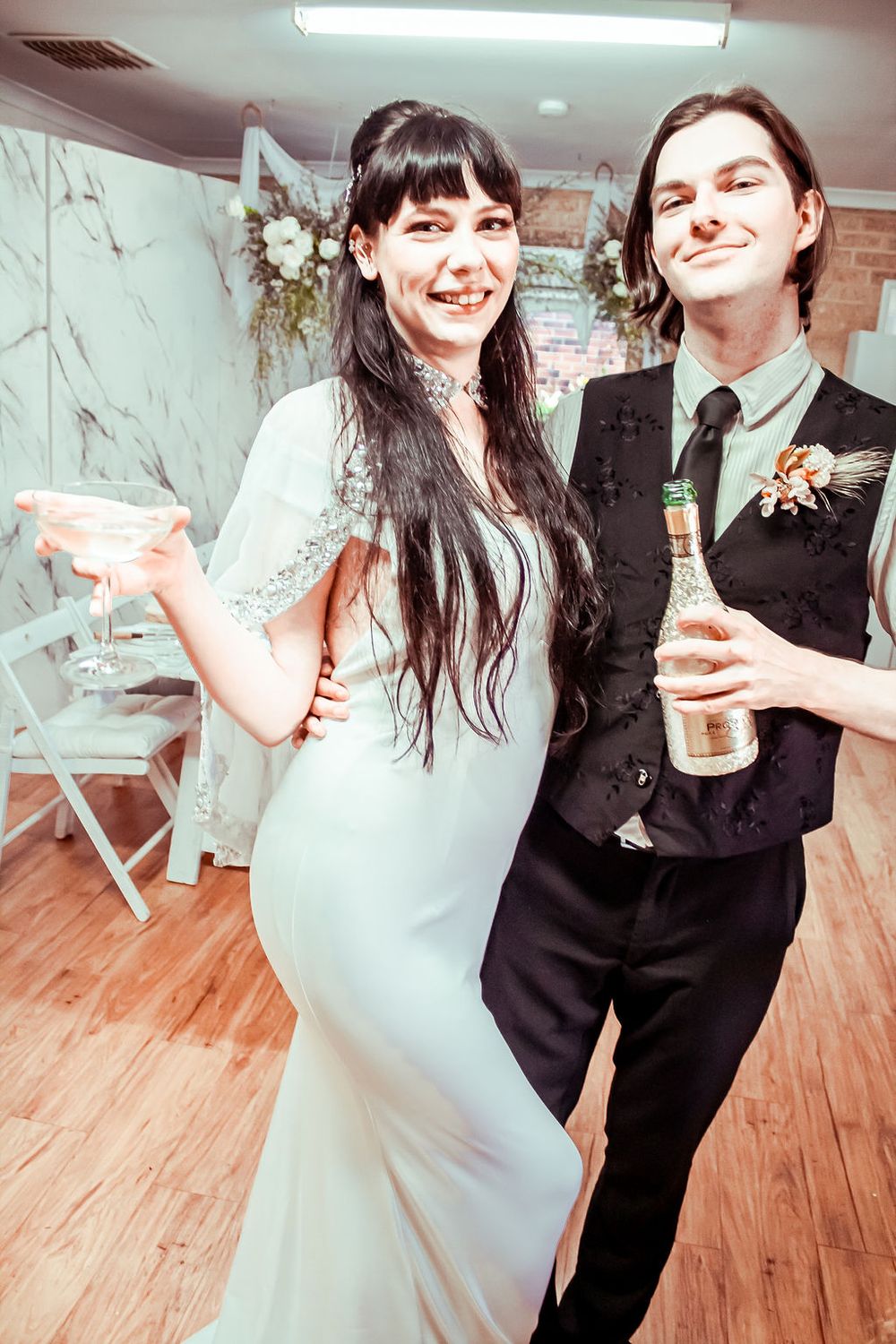 Newlywed couple celebrating at their wedding reception with martini glass and beer bottle in an indoor venue.