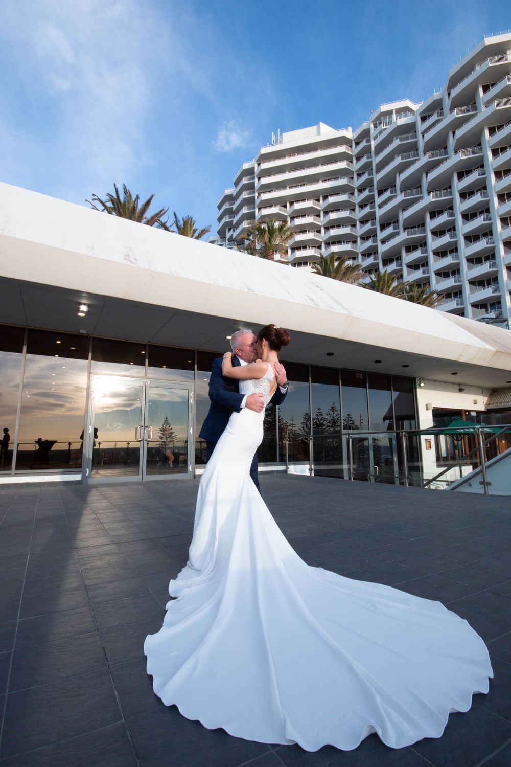 Wedding couple embracing in front of modern high-rise hotel with stunning white flowing bridal gown train.