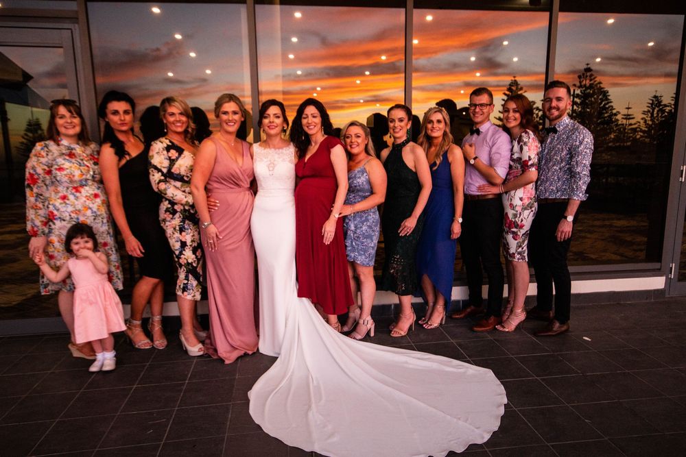 A wedding celebration group poses together at sunset with a bride in a white gown with train in the foreground.
