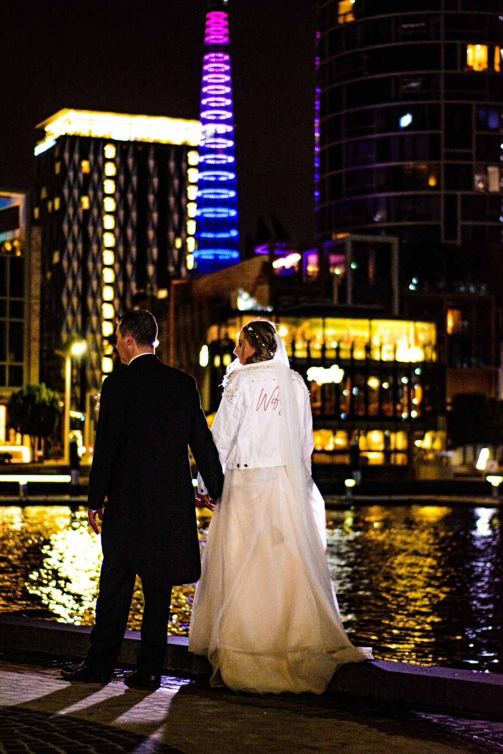 Couple admires city skyline with illuminated buildings reflecting on waterfront at night.