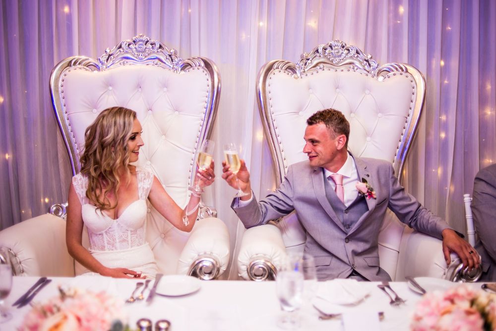 A couple toasts champagne while seated in ornate white throne chairs at their wedding reception with pink floral decor.