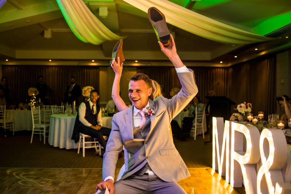 Person in grey suit celebrates on dance floor at wedding reception with illuminated MR sign.