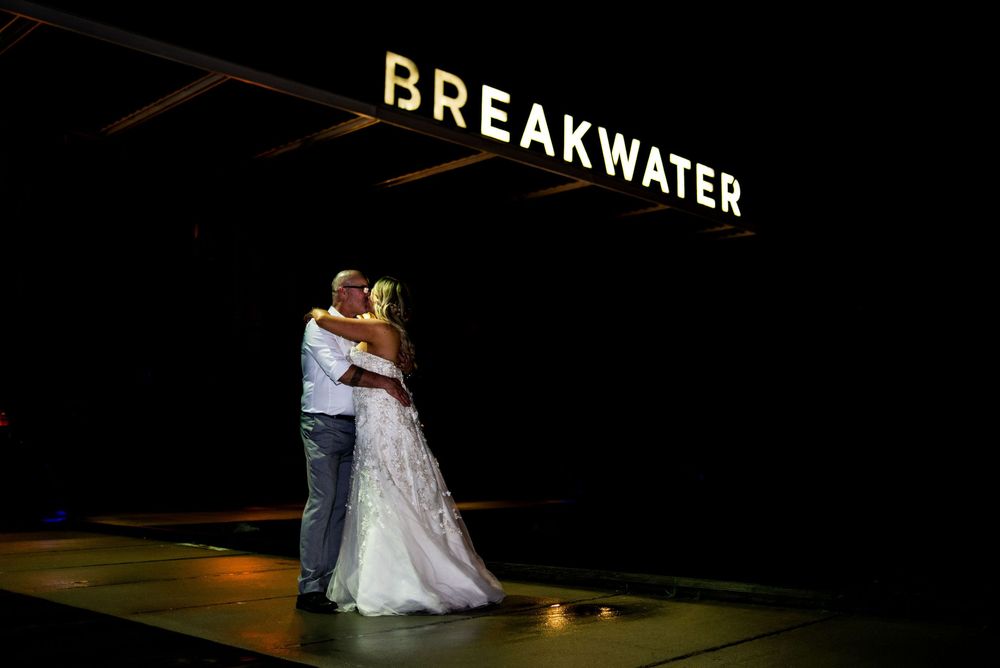 A couple embraces during their first dance under illuminated Breakwater signage at a nighttime wedding reception.