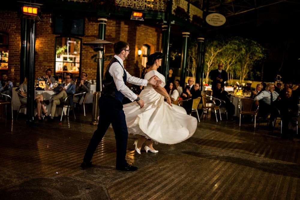 Couple performs their first dance on a wooden floor while guests watch in an industrial-style venue.