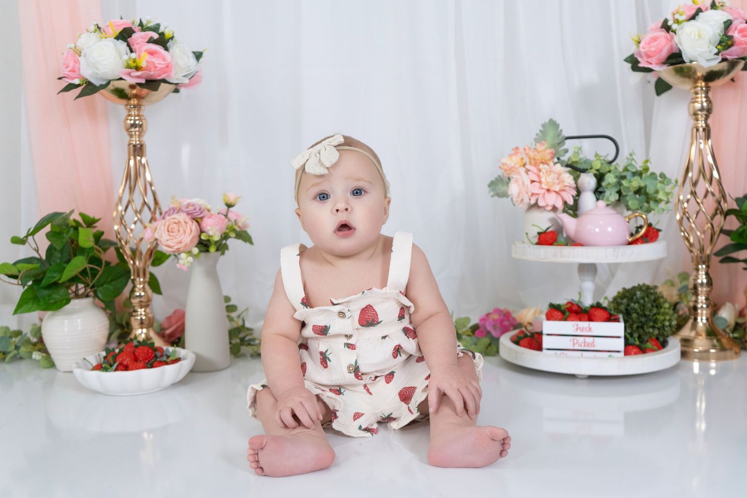 A baby in a floral dress sits on white backdrop surrounded by elegant flower arrangements and fresh strawberries baby milestone photographer