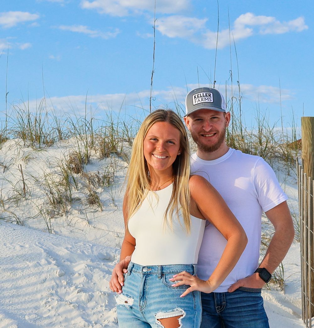 A couple stands together smiling on white sand dunes on a sunny day with blue skies.
