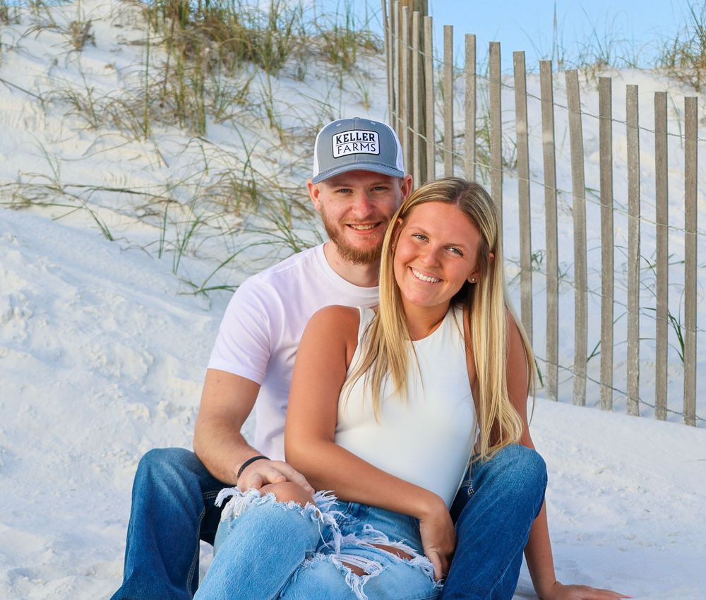 A couple sitting together on white sand with a beach fence in the background on a sunny day.