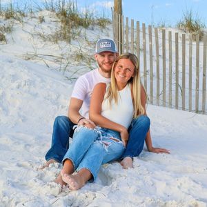 A couple sits together on white sand beach by wooden fence and sea grass on a sunny day.