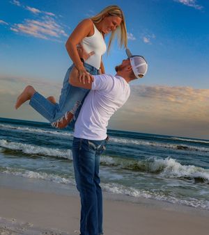 A romantic couple shares a playful moment on the beach at sunset as waves crash on the shore in the background.