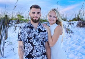 A couple poses together on a white sandy beach with sea grass, wearing a navy floral shirt and white sleeveless top.