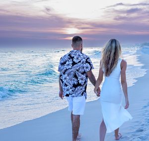 A couple walks hand in hand along a white sandy beach during a colorful sunset.