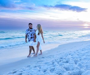A couple walks together on a white sandy beach at sunset with turquoise waters and pink-purple sky in the background.
