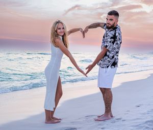 Two people make a heart shape with their hands while standing on a white sandy beach at sunset by the ocean.