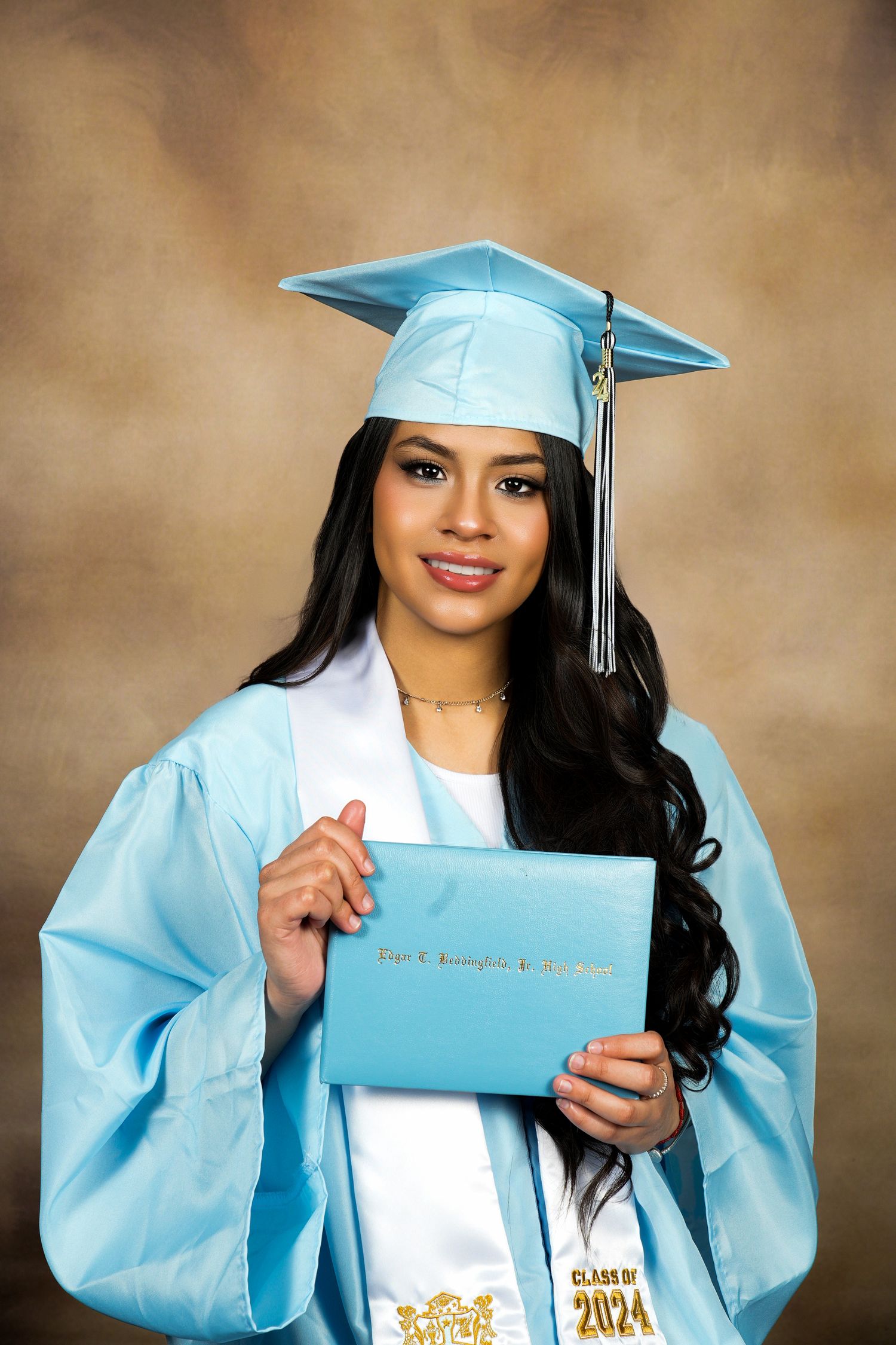 Graduate in light blue cap and gown holds diploma while posing for portrait against brown backdrop.