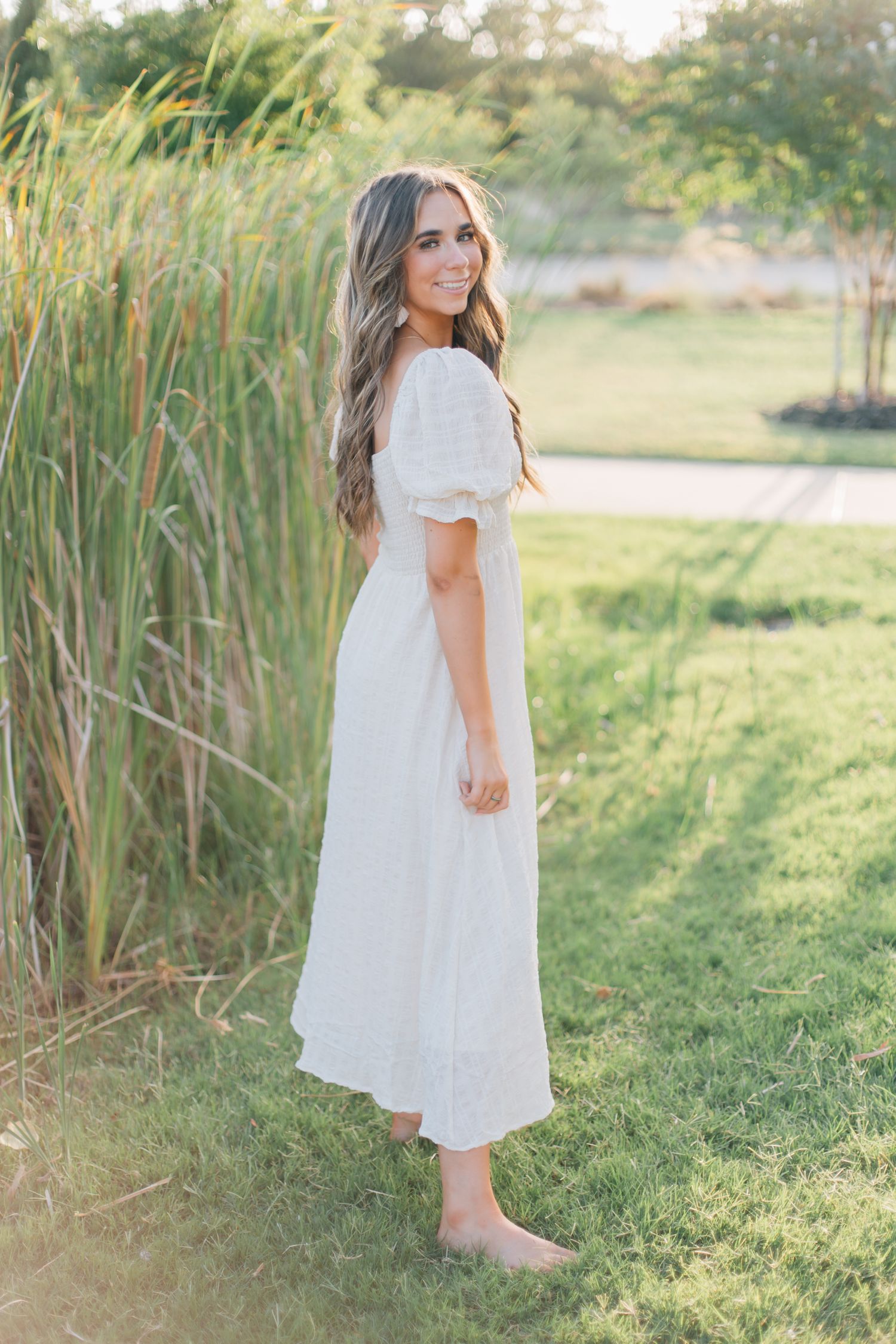 Full-length portrait of a woman in a flowing white dress posed against tall grass at sunset.